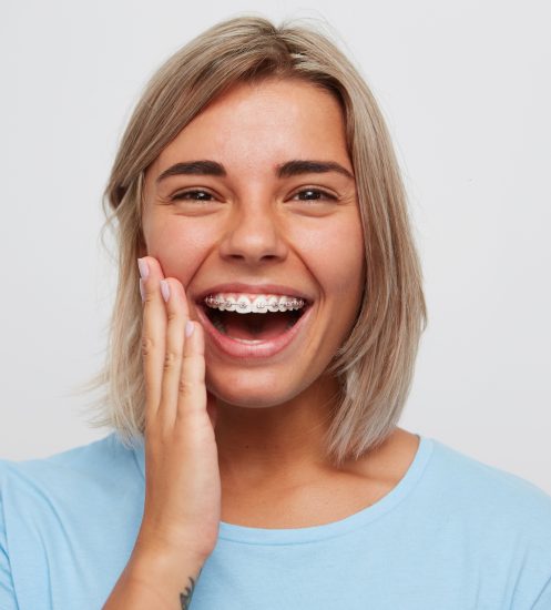 Cheerful beautiful young woman with blonde hair and braces on teeth laughing and touching her face by hand isolated over white background